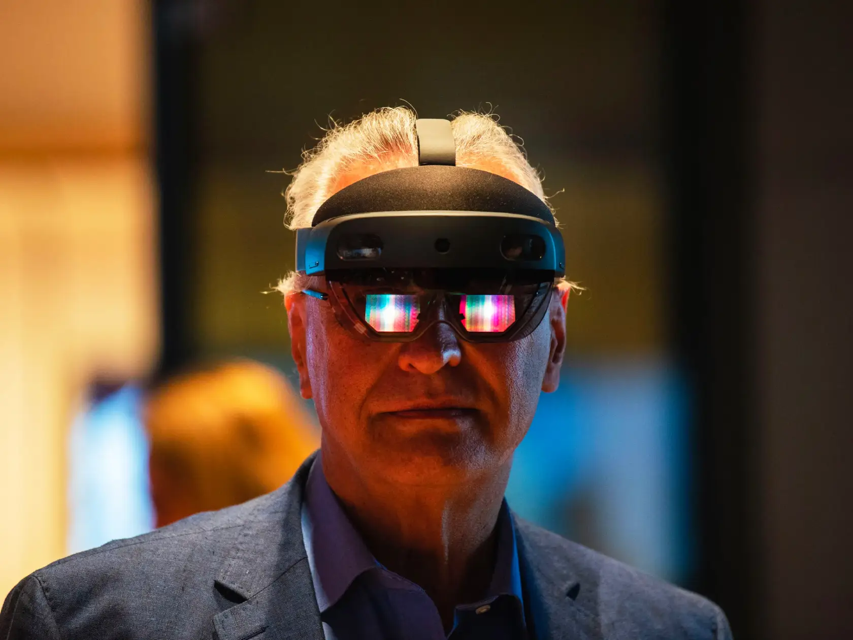 An old man in a blue suit looking into the camera wearing a sleek black augmented reality headset called a Microsoft Hololens.