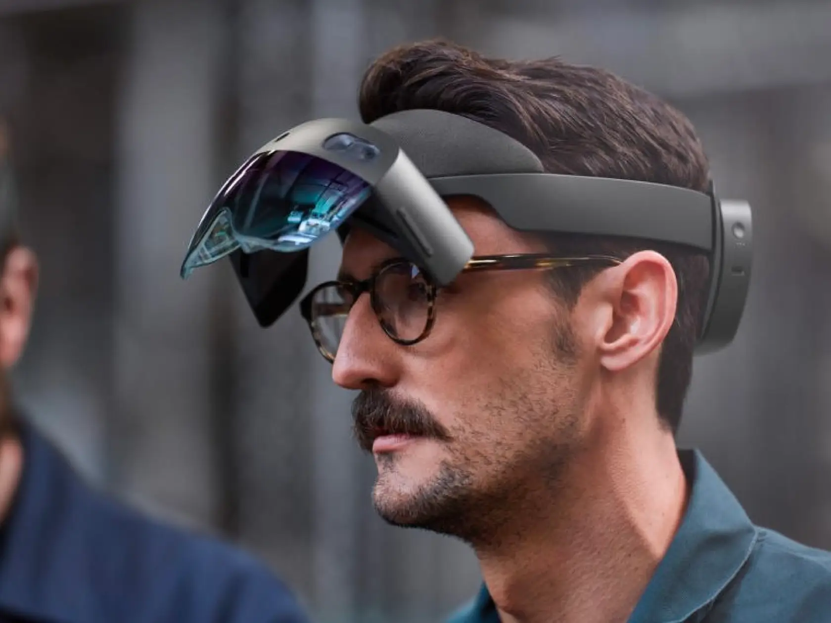 A man with a moustache and glasses wearing a black augmented reality headset with the visor lifted up